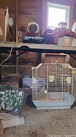 Photo showing resin metal birdcage on garage floor with bird accessories inside, nearby green bag, and wooden wall background.