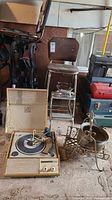 Overview of lot showing folding step stool chair, record player, metal rack, planter and iron scattered on floor in garage