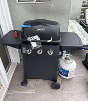 Full front view of the black Dyna-Glo gas grill including propane tank and side shelves on patio area.