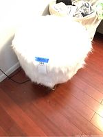 Angled side view of a square footrest stool covered in white faux fur upholstery, standing on a hardwood floor near a wall outlet.