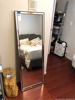 Large rectangular full-length mirror leaning against a wall next to a small wooden stump and suitcase, showing reflective surface and soft lighting.