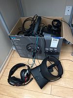 Box containing various cables and gaming electronics with Oculus Rift headset and gaming headset placed around it on a wooden floor.