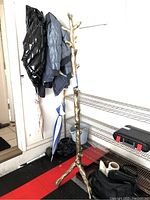 Full view of gold-tone tree trunk coat rack standing approximately 72 inches tall with coats and umbrellas hung on it in a garage setting.