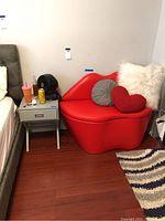 Red lips-shaped chair with three decorative pillows next to a small gray nightstand on hardwood floor.