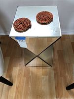 Top view of the mirrored end table with two orange and black woven coasters on top; shows reflective mirrored surface and smooth finish.