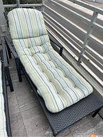 Outdoor wicker lounge chair with striped cushion showing wear and rip damage, placed on balcony space with railing and floor tiles.