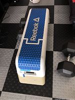 Top-down angled view of the Reebok weight bench showing textured blue padding with Reebok logo and white frame on exercise floor.