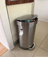 Photo of tall chrome trash can placed against wall and kitchen counter, showing lid and some trash inside.