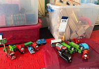 Overall view of various wooden Thomas and Friends trains and train cars arranged on a red surface near a storage bin containing wooden track pieces.