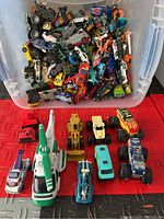 Top view of a plastic tub filled with numerous toy vehicles including cars, trucks, and motorcycles in a variety of colors.