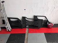 Wide photo showing three black metal rock sliders or side steps with perforated panels, vintage OEM style, placed side by side against a wall with a ladder and a bucket on the side.