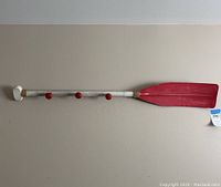 Front view of wooden oar mounted horizontally showing four round red pegs for hanging hats, painted white shaft and red blade end.