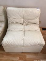 Front view of white leather armless chair with visible stitching and cushioned seat and backrest. Chair positioned against a wall on hardwood floor.