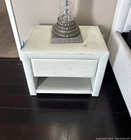 Front view of white leather bedside table with one drawer and open lower shelf, showing stains and dirt on top surface.
