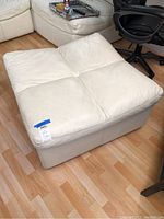 View of white leather square ottoman showing its tufted cushion top and boxy base on light wood floor with adjacent furniture.