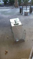 Photo of steel wire mesh animal trap placed on concrete floor, showing the top with a metal plate and handle for operation.