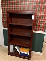 Full front view of the wooden bookcase showing three fixed shelves and various binder items on the bottom shelf (not included). Dark reddish-brown wood finish.