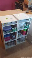 Front angled view of two white particle board shelving units side by side with various containers on shelves. Top surfaces partially visible with no tops included.