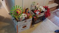 Photo of two cardboard boxes full of fabric faux florals and a colorful outdoor pinwheel decoration, positioned by a door and staircase.