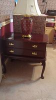 Antique style wooden cabinet table with two drawers, gold tone metal pulls, and a decorative lamp on top.