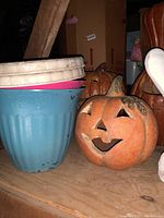 Photo showing ceramic pumpkins with smiling jack-o'-lantern faces alongside stacked plastic flower pots including blue, pink, and beige colors.