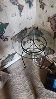 Full view of round glass top table on black metal stand placed in corner with map wallpaper background.