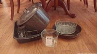 Shows aluminum speckled stock pot with lid, roasting pan, glass measuring pitcher, cut glass bowl, Pyrex bowl, and cast iron skillet on wood floor