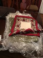 Two tablecloths, one olive green and one floral pattern with red border, stacked and partially wrapped in plastic on wooden table