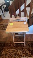 Full view of one oak tone solid wood TV table with folding top and legs, on carpeted floor near stairs.