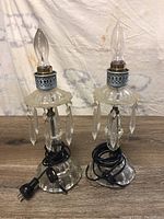 Pair of clear glass vintage-style lamps with hanging glass prisms, perforated metal collar, and flame-shaped bulbs, cords coiled on base.