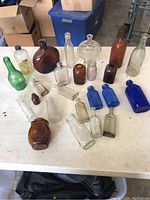 Full view of 20 antique glass bottles on table, various colors and shapes, showing worn but intact condition