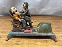 Side view of entire cast iron bank showing dentist leaning over patient in chair, foot pedal, and green coin bank slot on blue rectangular base with four feet.
