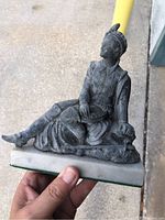 Photo of the figure front and side view showing seated person in detailed clothing and headdress on white marble base.