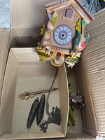 Birdhouse shaped cuckoo clock with carved leaves, bird, and face with Roman numerals inside a cardboard box. Pinecone weights, pendulum, and chains are detached and placed inside the box.