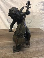 Full front view of bronze cherub playing cello statue on wooden surface, showing detail of entire figure and instrument.