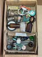 Top view of box containing various small collectible items including glass bottles, metal cups, ink pots, and vintage toy figurines.