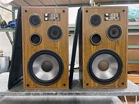 Front view of paired floor speakers showing wood grain cabinet, silver large woofer and three smaller speaker components, bass port, and overall wear.