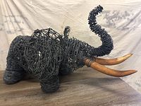 Side angled view of large wire elephant sculpture showing body shape, raised trunk, and two wooden tusks.