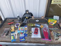 Wide view of the lot showing Batman figure, skateboard, brushes, toy cars, cardboard packaging of trade transformers, crayons, and other craft items on wooden surface.