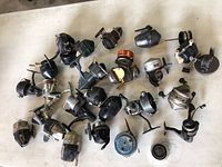 Overview of 23 vintage fishing reels arranged on a surface showing variety in styles, colors, and conditions.