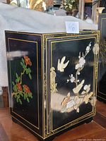 Front and side view of black lacquer cabinet showing floral and bird hand-painted design with gold trim