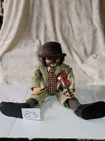 Photo shows a 22-inch clown hobo doll sitting with legs stretched out. The doll wears a brown hat, green shirt, plaid pants, and striped socks with black shoes. It holds a small red violin in one arm. The clown has a painted face with white makeup and exaggerated red lips and nose.