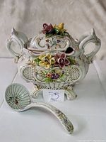 Front view of large ornate white pottery tureen with floral decorations on front, sides and lid, two large handles, and curved feet. Ladle is shown lying in front.