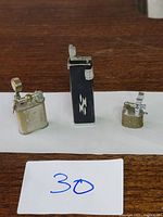 Three vintage miniature lighters displayed on a wooden surface: a silver lift arm lighter, a black lighter with white emblem, and a small lighter with mesh wrapping.