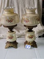Photo of a pair of midcentury side lamps with ceramic bases and shades decorated with floral patterns, standing on ornate brass bases against a neutral background.