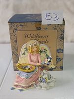 Figurine of a blonde angel dressed in pink with flowers, holding a basket and a small lamb, positioned in front of the original box.