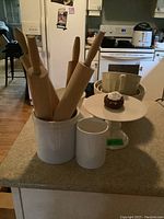 Kitchen counter with 4 wooden rolling pins inside white ceramic holder, white cylindrical holder, beige mixing bowls, and white ceramic cake stand with dessert.