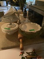 Kitchen counter with white ceramic cake stand with scalloped edges, clear glass cake stand, green glass cake stand, wooden rolling pin, and clear glass mixing and trifle bowls.
