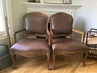 Pair of brown upholstered wood framed armchairs with carved details and nailhead trim shown from front side view on wooden floor.
