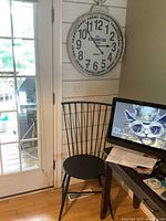Photo shows black spindle back chair next to a desk and Kensington wall clock hung on beige paneled wall near glass door.